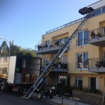 Vor einem gelben Mehrparteienhaus steht ein Umzugs-LKW. Ein Lastenaufzug ist am Haus aufgestellt und reicht bis zur Dachterrasse im dritten Stockwerk.